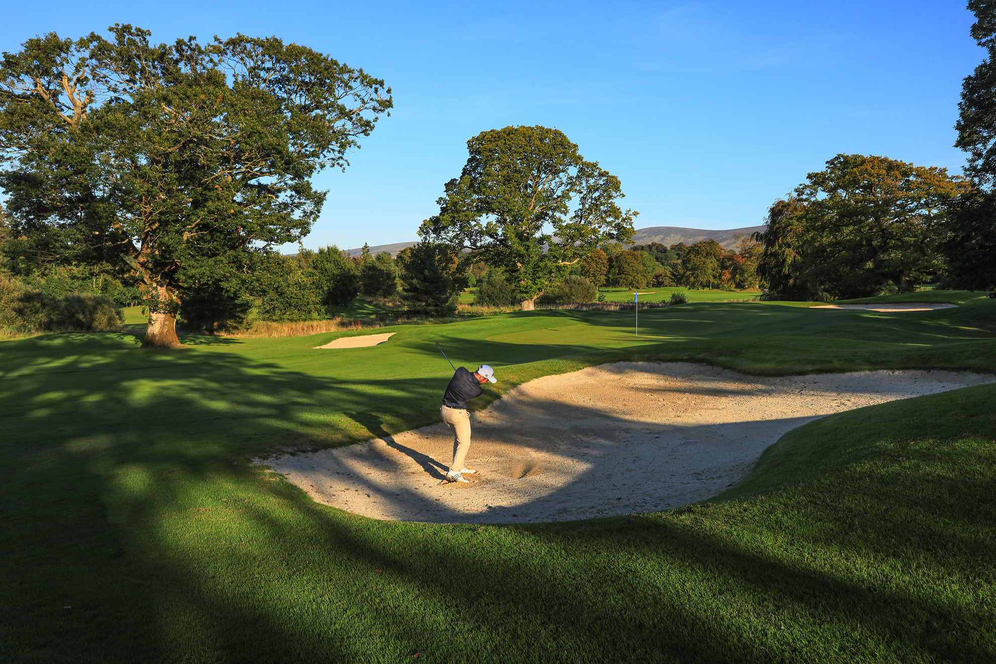 Gary Shaw Golf hitting a bunker shot onto a green with sunshine in the background. 